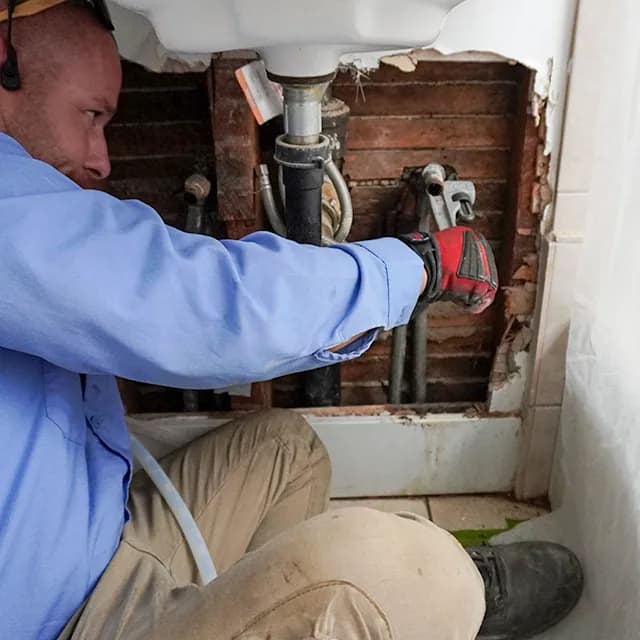 Technician repairing leaking drain plumbing beneath a bathroom sink